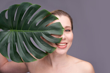 Caucasian female adult smiling behind monstera leaf on gray background.