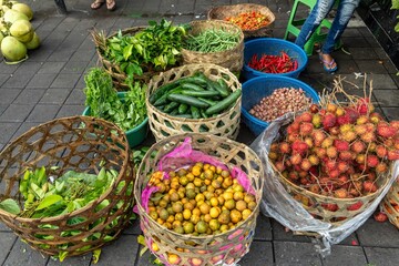 A street vendor displays fresh produce in woven baskets at a market in Ubud, Bali, Indonesia. The colorful array includes rambutan, oranges, cucumbers, and various greens.