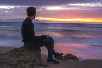 Man sitting on beach rocks looking at ocean during sunset