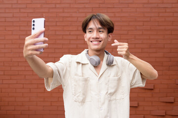 Non binary cute Asian young gay man using mobile phone against brick wall