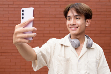 Non binary cute Asian young gay man using mobile phone against brick wall