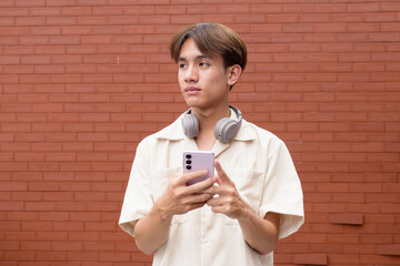 Non binary cute Asian young gay man using mobile phone against brick wall