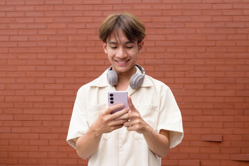 Non binary cute Asian young gay man using mobile phone against brick wall