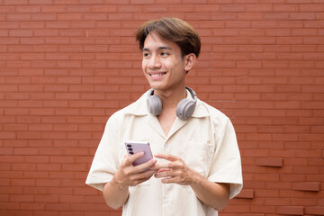 Non binary cute Asian young gay man using mobile phone against brick wall