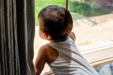 A curious young Asian baby looking out through a window in natural sunlight. A warm parenting...