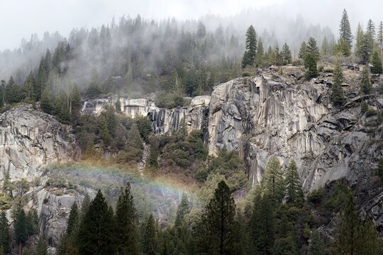 Misty mountain landscape with a rainbow arching over rocky cliffs and a dense forest