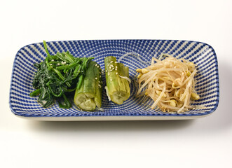 Rectangular blue and white patterned plate contains three types of Korean side dishes seasoned spinach, pickled cucumbers, and seasoned soybean sprouts, garnished with sesame seeds