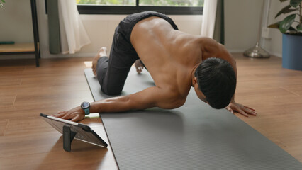 Asian man is exercising and stretching at home while watching a workout routine on his tablet.