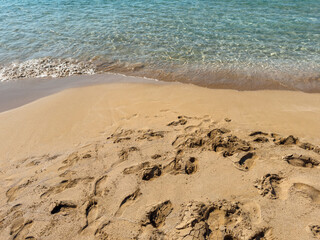 Golden sand beach with clear turquoise water and gentle waves washing ashore, featuring natural textures of sand, shallow sea and footprints under bright summer sun