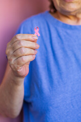 Close-up image of a senior woman in blue t-shirt gently holding a pink breast cancer awareness...