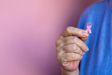 Close-up image of a senior woman in blue t-shirt gently holding a pink breast cancer awareness...