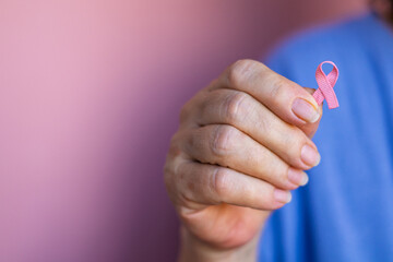 Close-up photo of an elderly woman in blue t-shirt gently holding a pink ribbon in her hand,...