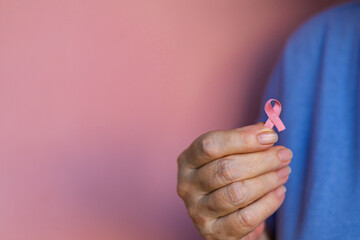 Close-up of an elderly woman in blue t-shirt gently holding a pink ribbon in her hand, symbolizing...