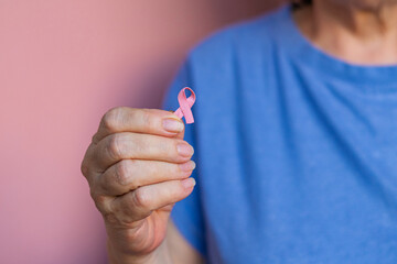 Close-up of an elderly woman in blue t-shirt gently holding a pink ribbon in her hand, symbolizing...
