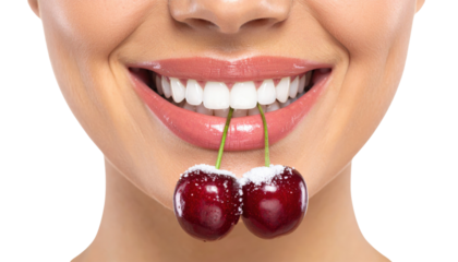 A close-up of a smiling mouth holding two fresh cherries with green stems between the teeth.