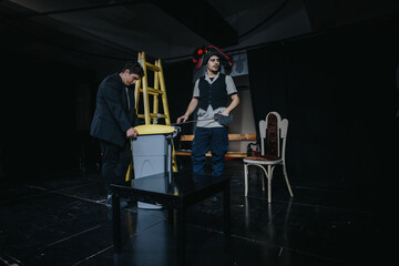 An artistic theatrical performance featuring two actors in costume on a dramatic stage setup with...