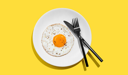 Fried eggs with spicy and black cutlery on a white plate on a bright yellow background with shadow.