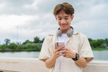 Asian non binary tourist person with headphones relaxing at park near lake using mobile phone