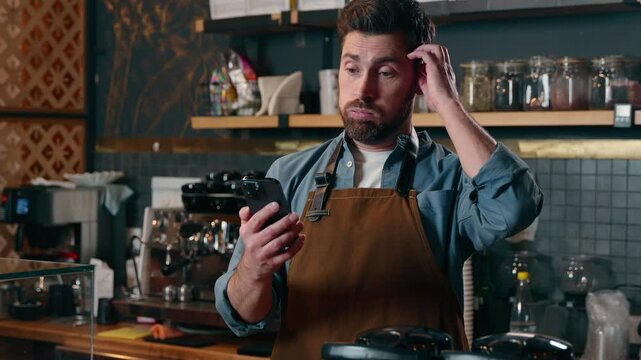 Frustrated confused Caucasian barista man look screen mobile phone in cafeteria male small business owner upset bartender using smartphone waiter guy money loss difficulty online trouble failure cafe