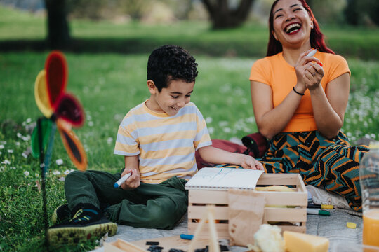A Filipino mother and her son share laughter during a creative activity in a park. The warm setting includes colorful elements and a joyful atmosphere, showcasing family bonding and outdoor happiness.