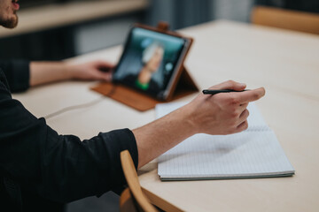A young male actively engages in an online learning session, taking notes on a paper notebook while using a digital device for virtual interactions.