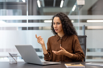 A businesswoman has a video call in a modern office, actively gesturing as she speaks into her laptop.