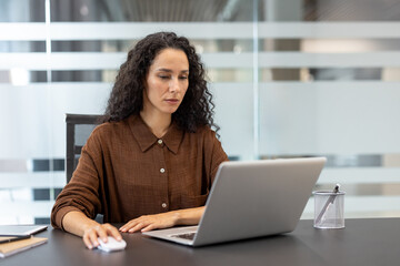 A woman with curly hair works on a laptop in an office setting, focusing intently on her computer...