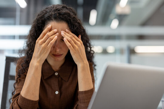 A woman stressed out at her desk, touching her head, looking down, with a laptop in front of her in an office setting.