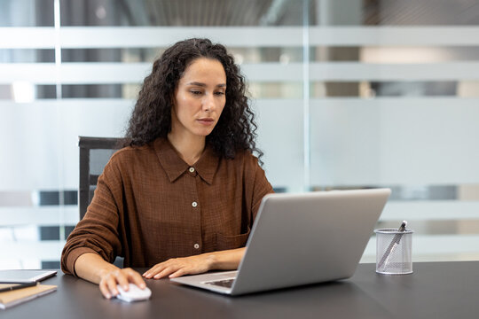 A woman with curly hair works on a laptop in an office setting, focusing intently on her computer screen.