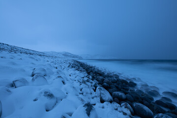Dragon's eggs beach in Teriberka. Snow blankets a rocky shoreline, meeting the calm sea under a twilight sky. Severe North landscape