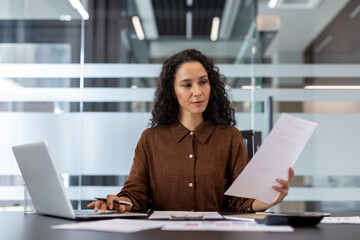 A businesswoman works in a modern office, analyzing documents while using a laptop, emphasizing...