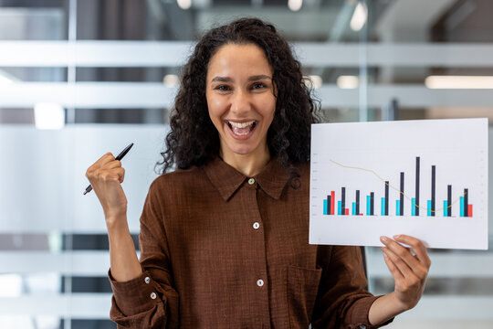 A happy woman in a brown shirt holding a chart with a graph and a pen, celebrating business success.