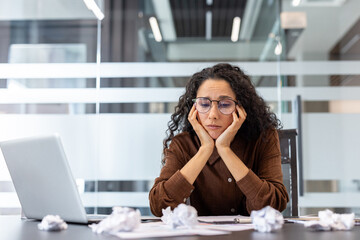 A woman in glasses looks despondent, resting her head on her hands at her desk, surrounded by...