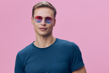 Man wearing pink sunglasses and blue t-shirt posing against a pink background, lifestyle image,...