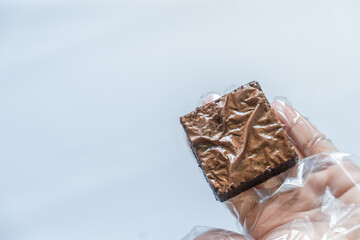 Gloved Hand Holding Brownie in Right Foreground on White Background