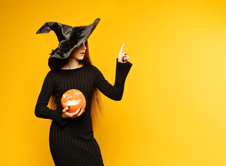 Young woman wearing black dress and hat, holds pumpkin jack o lantern on yellow background. Concept...