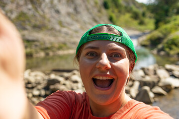 Energetic young woman in a green cap and orange shirt taking a joyful selfie by a mountain stream....