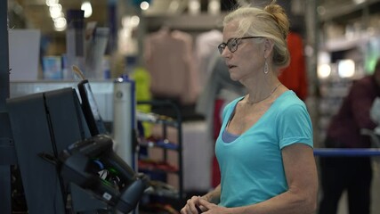 An unhappy, frustrated, mature woman stands at the self checkout, looking confused as she prepares to finalize her purchase with a credit card.