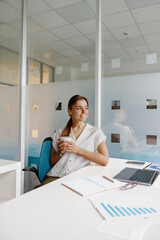 A thoughtful woman is seated in a modern office, carefully holding a cup amidst various work...