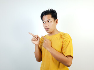 Young man in a yellow shirt pointing sideways with both hands, expressing curiosity and interest,...