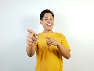 A cheerful young Asian man in a casual yellow shirt pointing forward, smiling joyfully on a white...