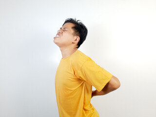 A young Asian man in a yellow shirt struggles with back pain, expressing discomfort against a white background. The image emphasizes health, pain, and physical strain concepts.