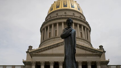 A prominent statue stands in front of the West Virginia State Capitol, showcasing its grand architecture and golden dome under an overcast sky.