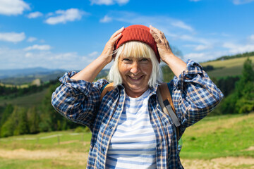 A cheerful elderly woman in a red beanie and plaid shirt standing in a picturesque mountain landscape. She smiling joyfully, enjoying the fresh air and nature during her hiking adventure. The clear