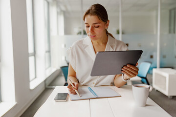 A professional woman engrossed in her work, utilizing a tablet and a notepad in a bright, modern...