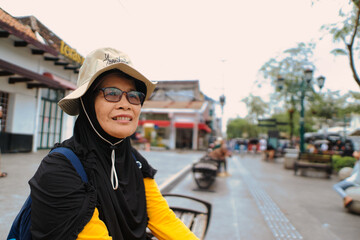 A portrait of a smiling elderly female tourist wearing a hijab, standing on the bustling Malioboro Street, evokes the cheerful and vibrant atmosphere of urban travel.