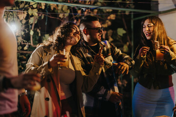 A group of cheerful friends are enjoying an evening together outdoors, holding drinks, laughing, and having a great joyful time at a celebration under soft lighting.