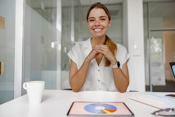 A cheerful woman sits in a vibrant office, ready for an important meeting with a tablet and a coffee
