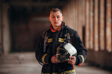 Holding helmet. Firefighter in professional equipment is indoors in abandoned building
