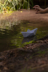 A person is throwing a delicate paper boat into a serene body of water, creating ripples in the...